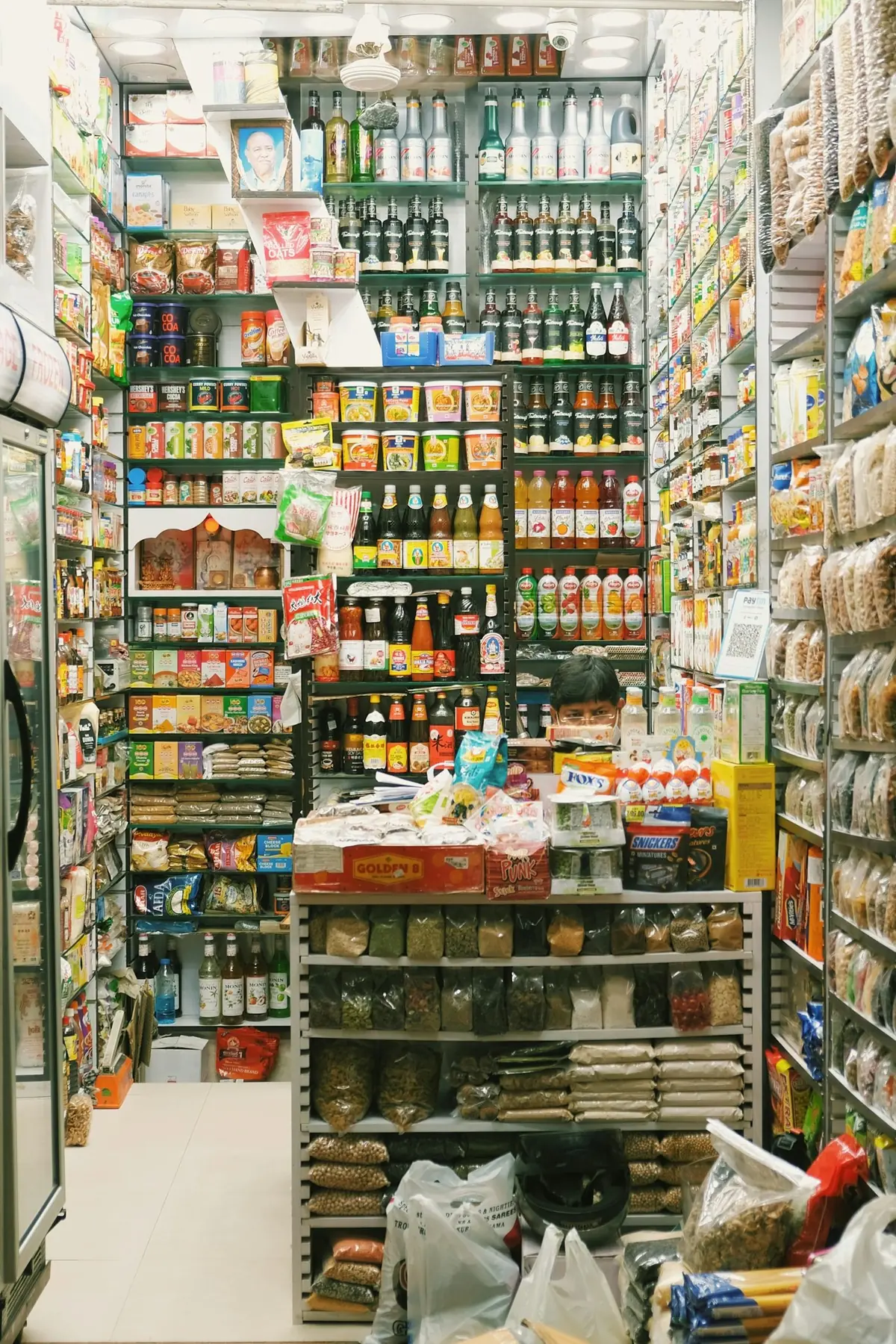 Pequena mercearia cheia de produtos embalados, com o comerciante atrás do balcão
